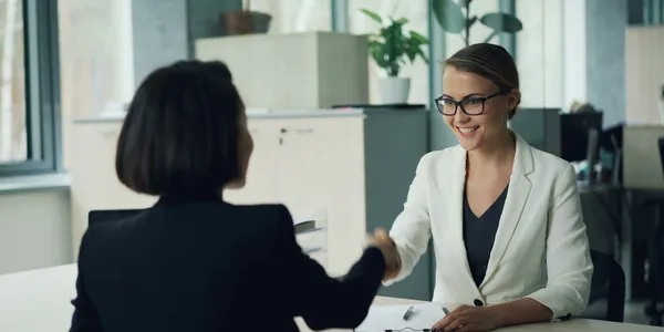 Two women shaking hands across a desk in an office