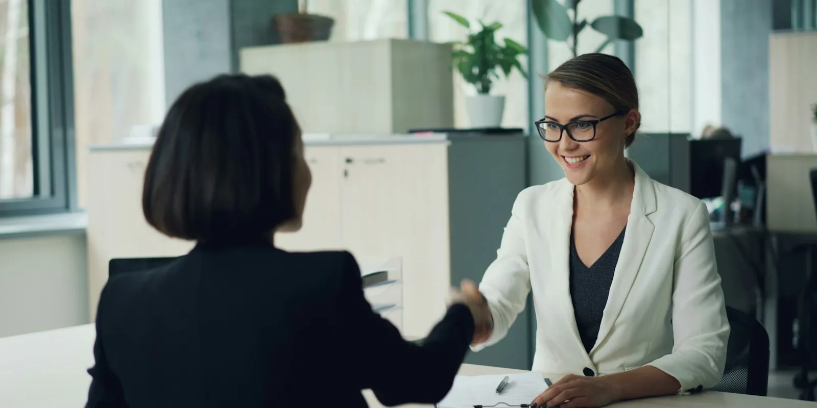 Two women shaking hands across a desk in an office